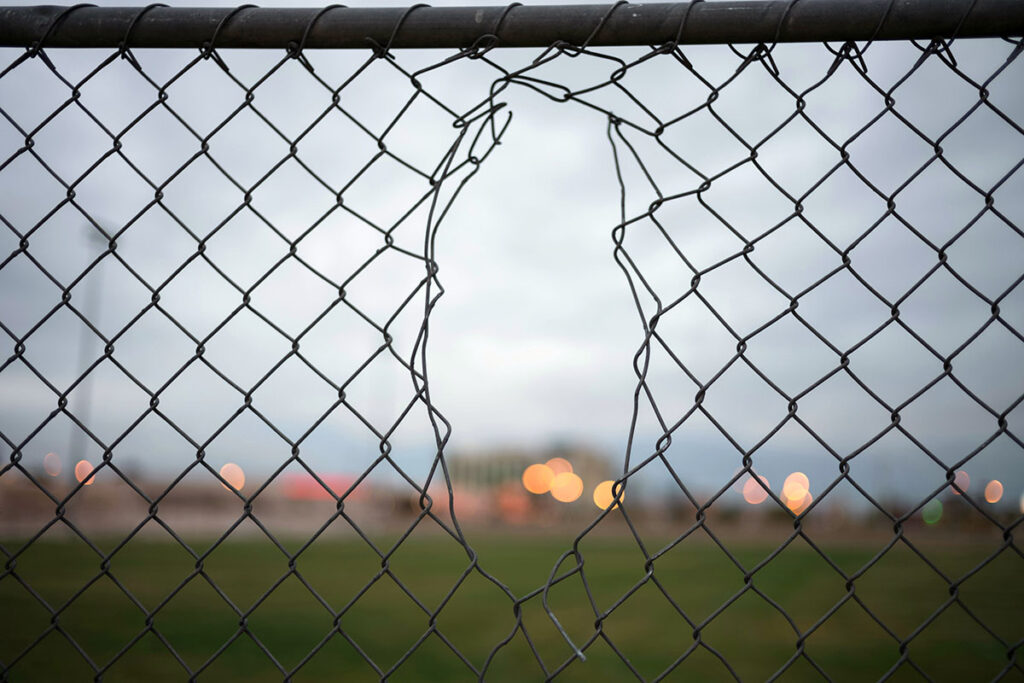 Close-up of a large, jagged hole cut into a wire chain-link fence, revealing a blurred field and distant lights in the background.