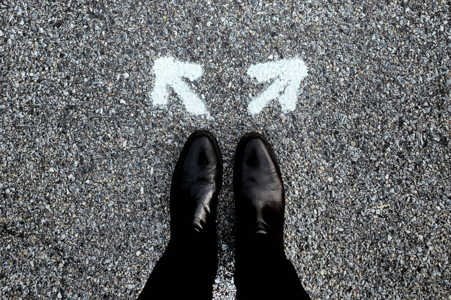 Close-up of a person's shoes standing on pavement with two arrows painted on the ground pointing in different directions