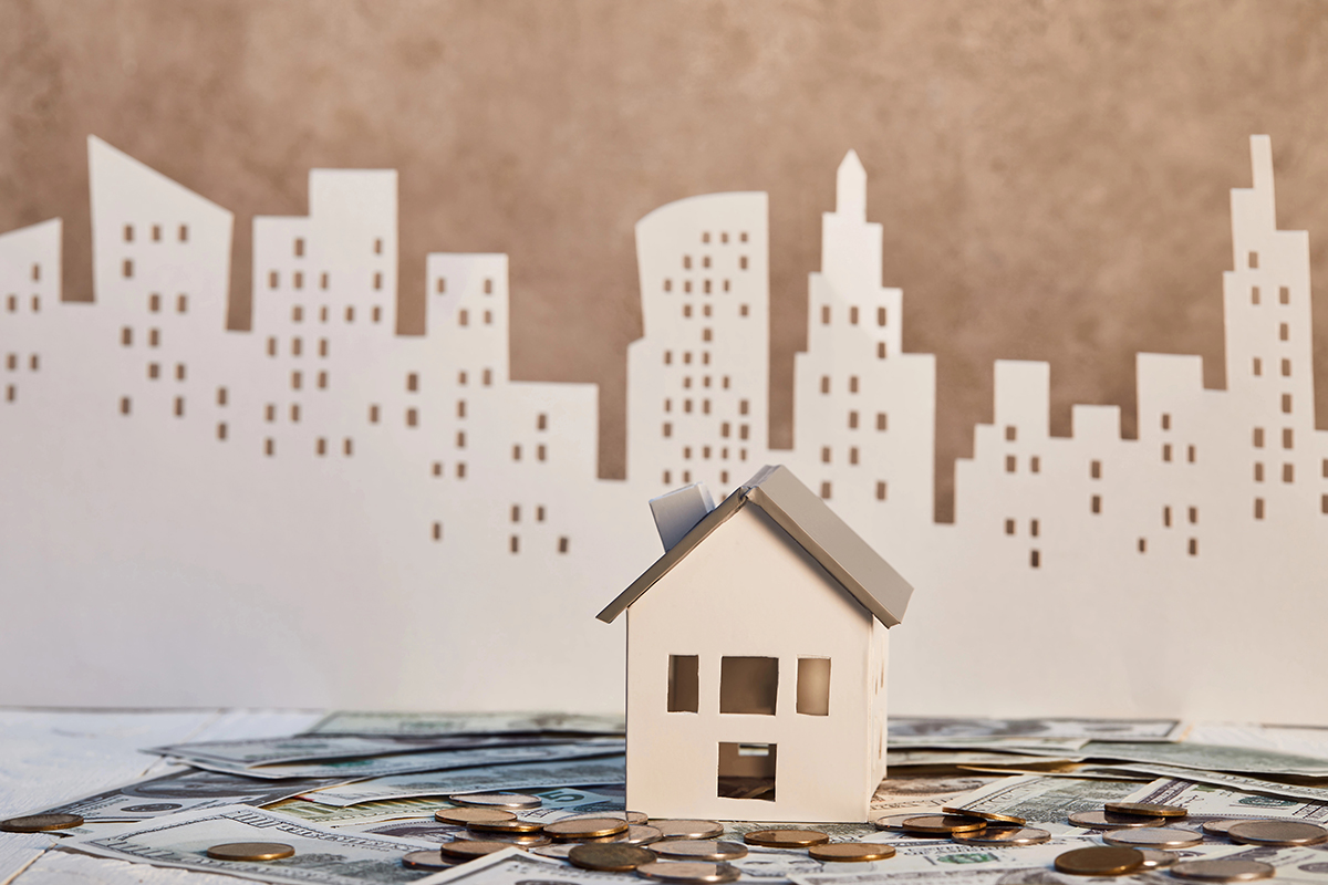 A miniature white paper model house with a grey roof sitting on a bed of US dollar bills and coins, with a paper city skyline cutout in the background.