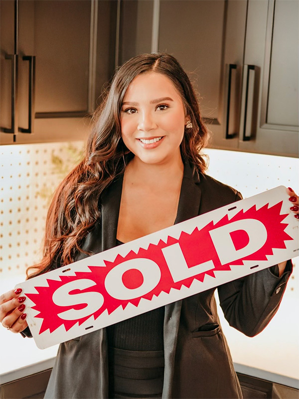 A smiling woman in a black blazer holds a red and white "SOLD" sign in a modern kitchen setting.