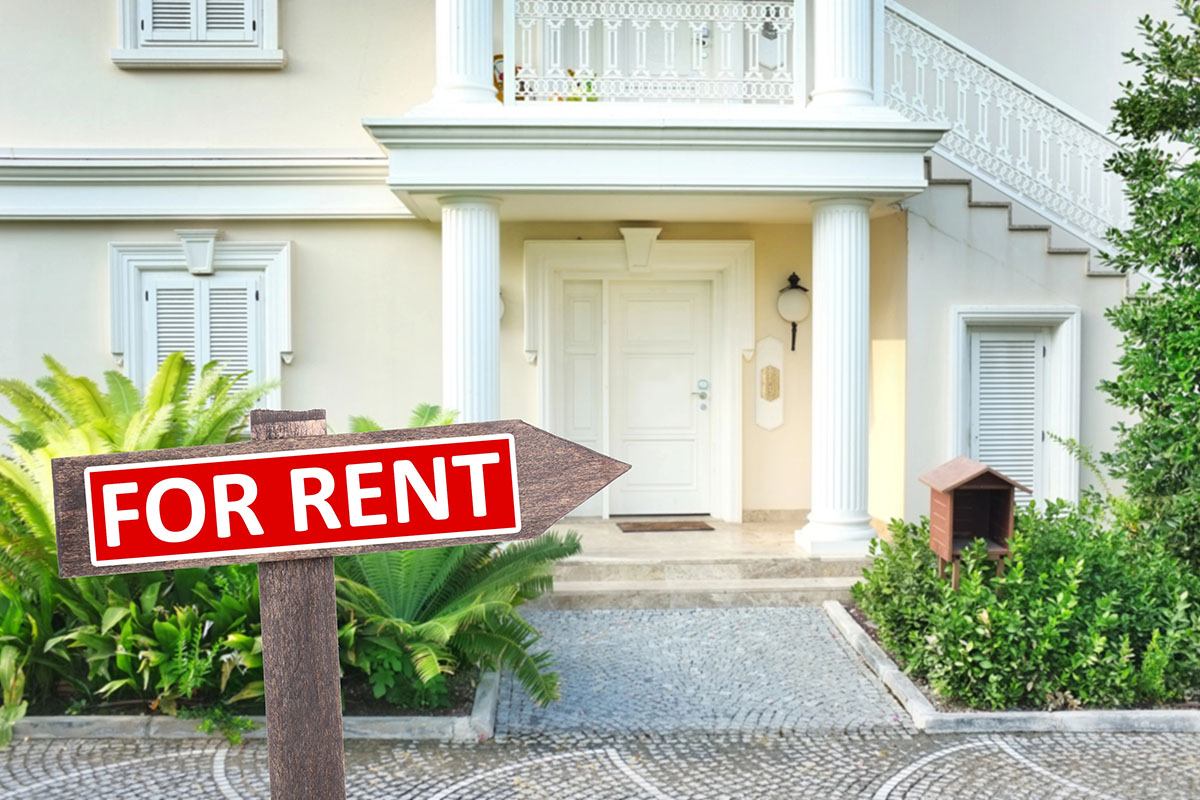 Front view of a house with columns, steps, and a "FOR RENT" sign in the foreground, surrounded by greenery and a stone pathway.
