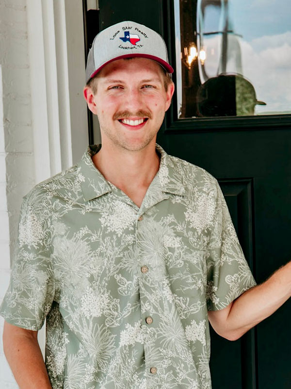A young man with a mustache stands smiling in front of a door. He wears a green floral short-sleeve shirt and a gray baseball cap with a Texas flag logo. The background includes part of a brick wall and a sunny sky.
