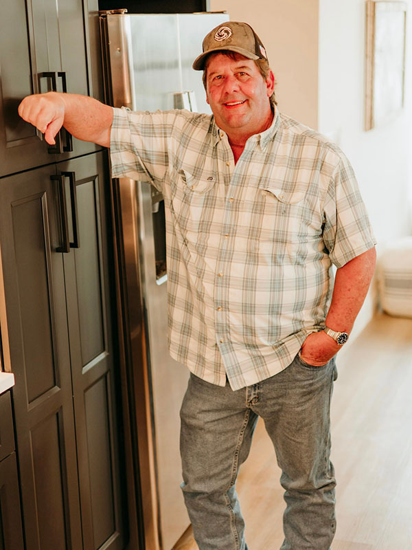 A smiling man in a plaid shirt and cap stands next to a refrigerator, casually leaning against a kitchen cabinet with his left hand on his hip.