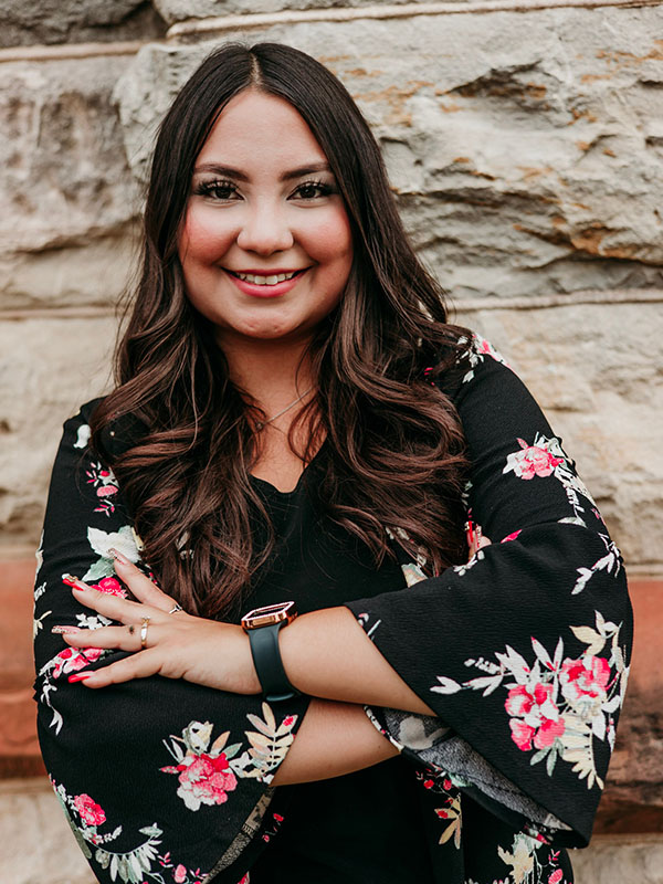 A woman with long, dark hair smiles confidently while standing with her arms crossed. She wears a black top adorned with floral patterns and is positioned in front of a stone wall.