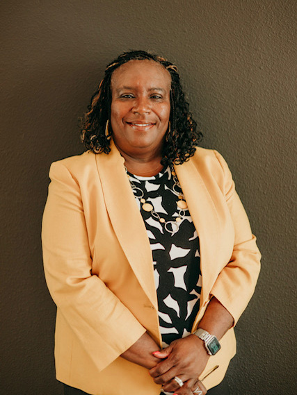 A smiling woman stands confidently against a dark wall, wearing a light yellow blazer over a black and white patterned top. She has curly hair and accessorizes with jewelry, including a watch.