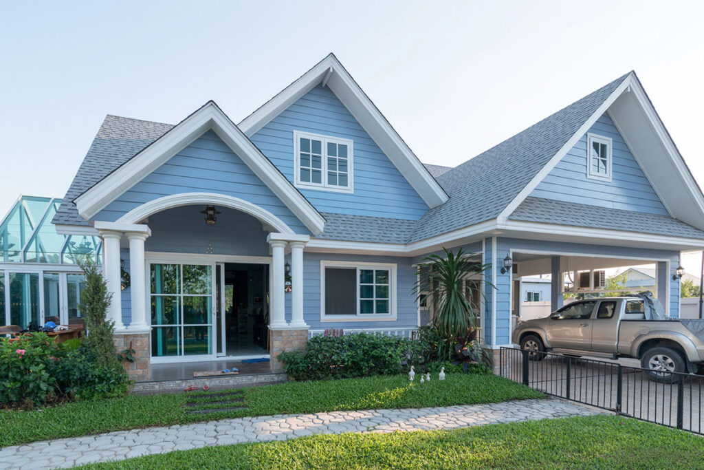 A light blue house with a gabled roof, white columns at the entrance, and landscaped greenery in the foreground. A silver pickup truck is parked under a carport on the right.