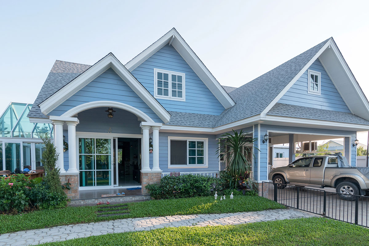 A light blue house with a gabled roof, white columns at the entrance, and landscaped greenery in the foreground. A silver pickup truck is parked under a carport on the right.