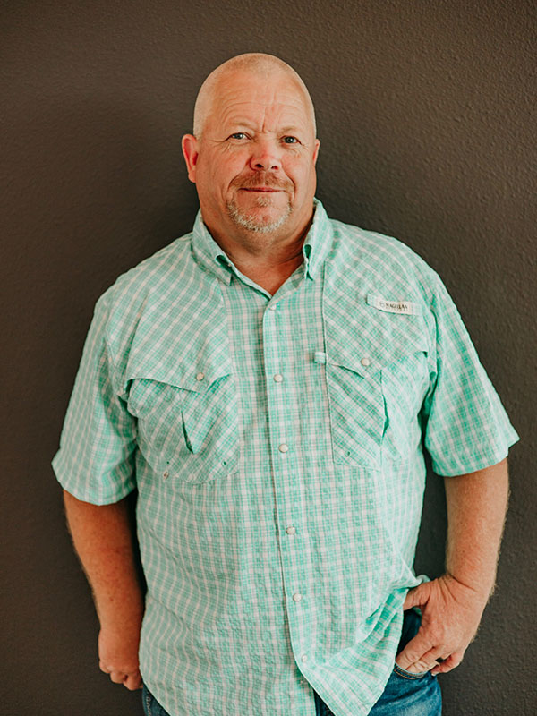 A man with a bald head and a light blue plaid shirt stands against a dark wall, smiling confidently with his hands on his hips.