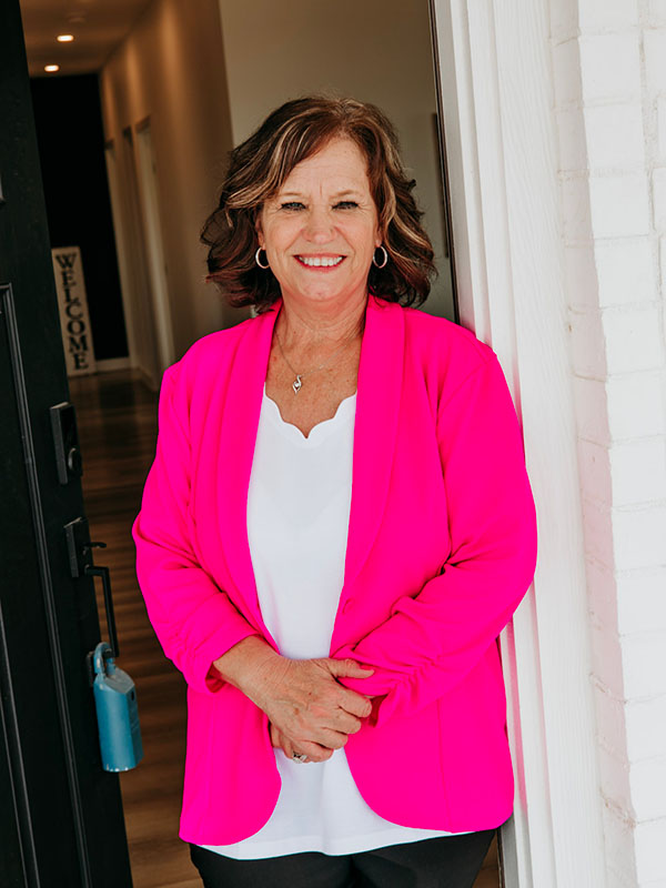 A woman with shoulder-length brown hair smiles while standing by an open door, wearing a bright pink blazer over a white top. She has hoop earrings and her arms are crossed in front of her. The background shows a well-lit hallway with a "WELCOME" sign.