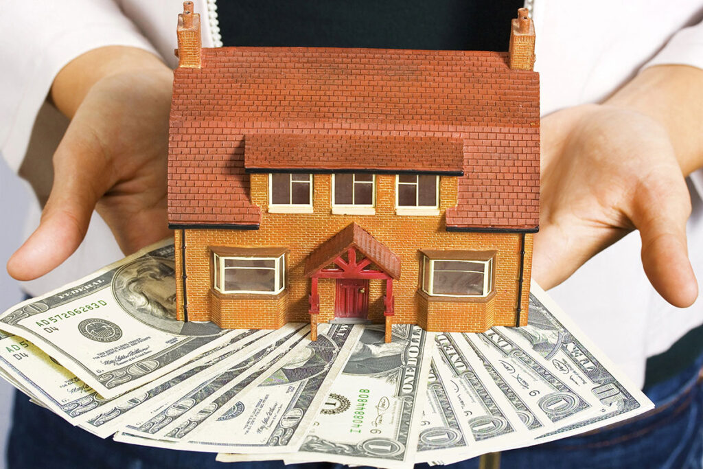 A miniature house is held above a fan of hundred-dollar bills, symbolizing real estate investment or home ownership.