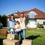 A happy family stands in front of their new home, with the parents holding house keys and the daughter hugging a teddy bear. Moving boxes are placed on the grass, and a van is parked on the side. The sky is clear and blue, and the house features a red roof and green landscaping.