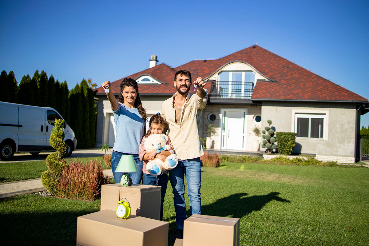 A happy family stands in front of their new home, with the parents holding house keys and the daughter hugging a teddy bear. Moving boxes are placed on the grass, and a van is parked on the side. The sky is clear and blue, and the house features a red roof and green landscaping.
