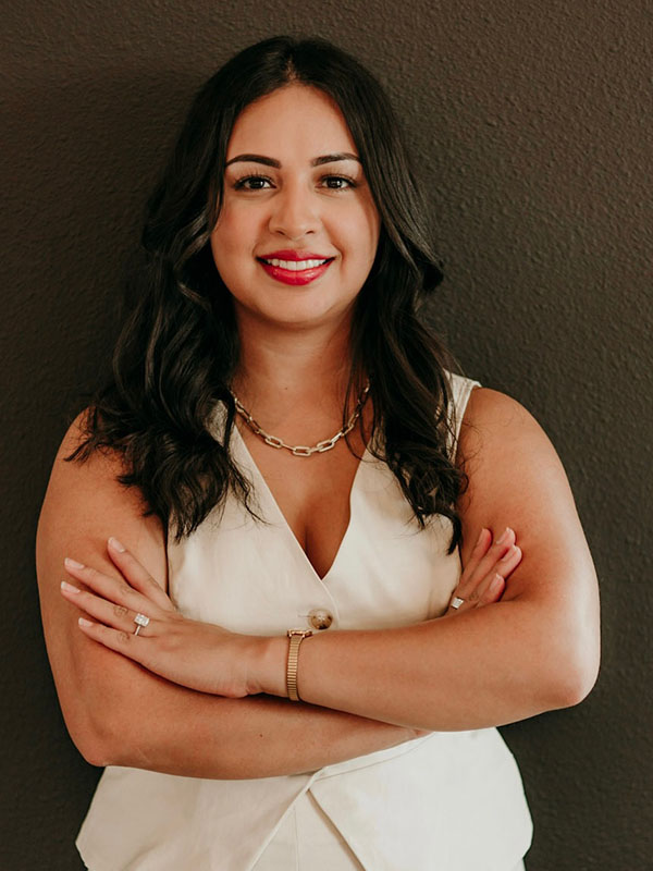 A woman with long, wavy dark hair and a warm smile stands confidently with her arms crossed. She is wearing a white sleeveless top and layered jewelry, standing in front of a dark wall.