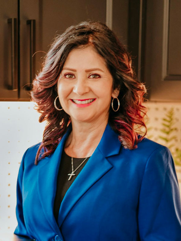 A woman with long, wavy hair featuring reddish highlights smiles warmly while wearing a blue blazer and silver hoop earrings, set against a modern kitchen background.