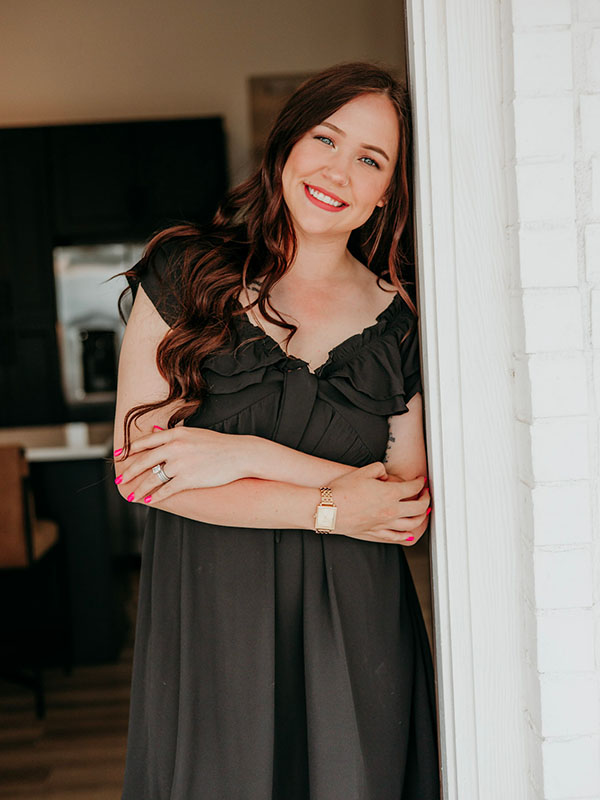 A woman with long, wavy brown hair and a bright smile leans against a white wall, wearing a black dress with ruffled details. She has her arms crossed and is accessorized with a gold watch, against a softly blurred background of a stylish kitchen.