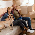 A couple sits on a couch in a room with unpacked cardboard boxes, smiling at the camera. They are accompanied by a yellow Labrador dog resting beside them. The woman wears a denim jacket and has her hair pulled back with a bow, while the man is dressed in a plaid shirt and a green t-shirt.