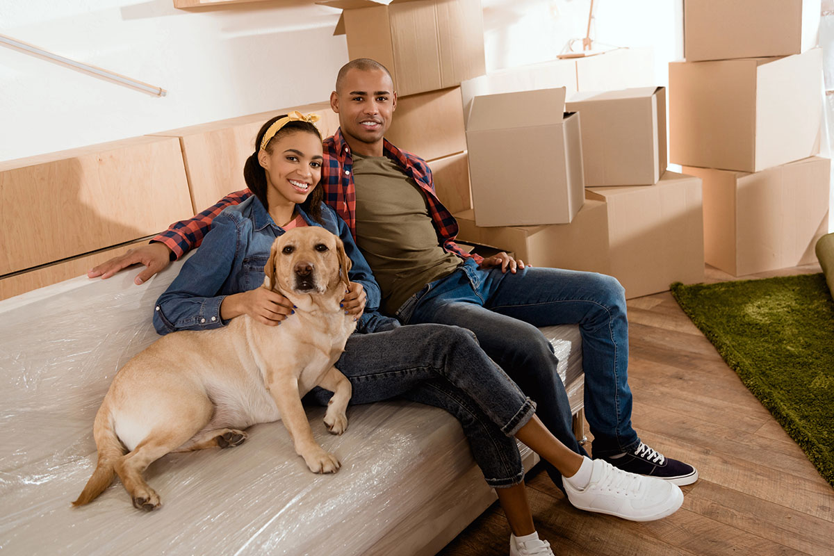 A couple sits on a couch in a room with unpacked cardboard boxes, smiling at the camera. They are accompanied by a yellow Labrador dog resting beside them. The woman wears a denim jacket and has her hair pulled back with a bow, while the man is dressed in a plaid shirt and a green t-shirt.