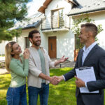 A couple smiles and engages in conversation while shaking hands with a real estate agent outside a modern home. The agent holds a clipboard. Lush greenery surrounds the scene.