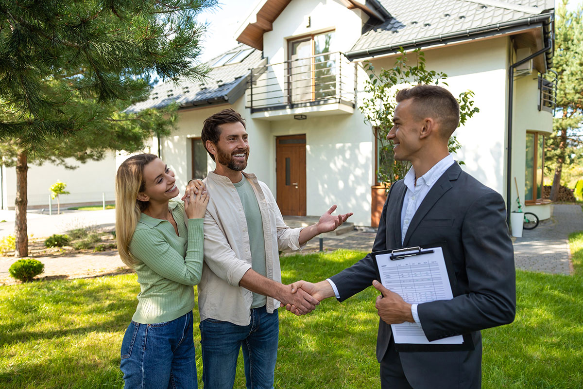 A couple smiles and engages in conversation while shaking hands with a real estate agent outside a modern home. The agent holds a clipboard. Lush greenery surrounds the scene.