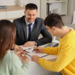 A professional man in a suit sits at a desk, smiling as he hands keys to a woman with long hair, who is wearing a green sweater. A young man in a yellow sweatshirt is looking at a document while holding a pen. A laptop is placed on the desk, and they appear to be discussing a transaction.