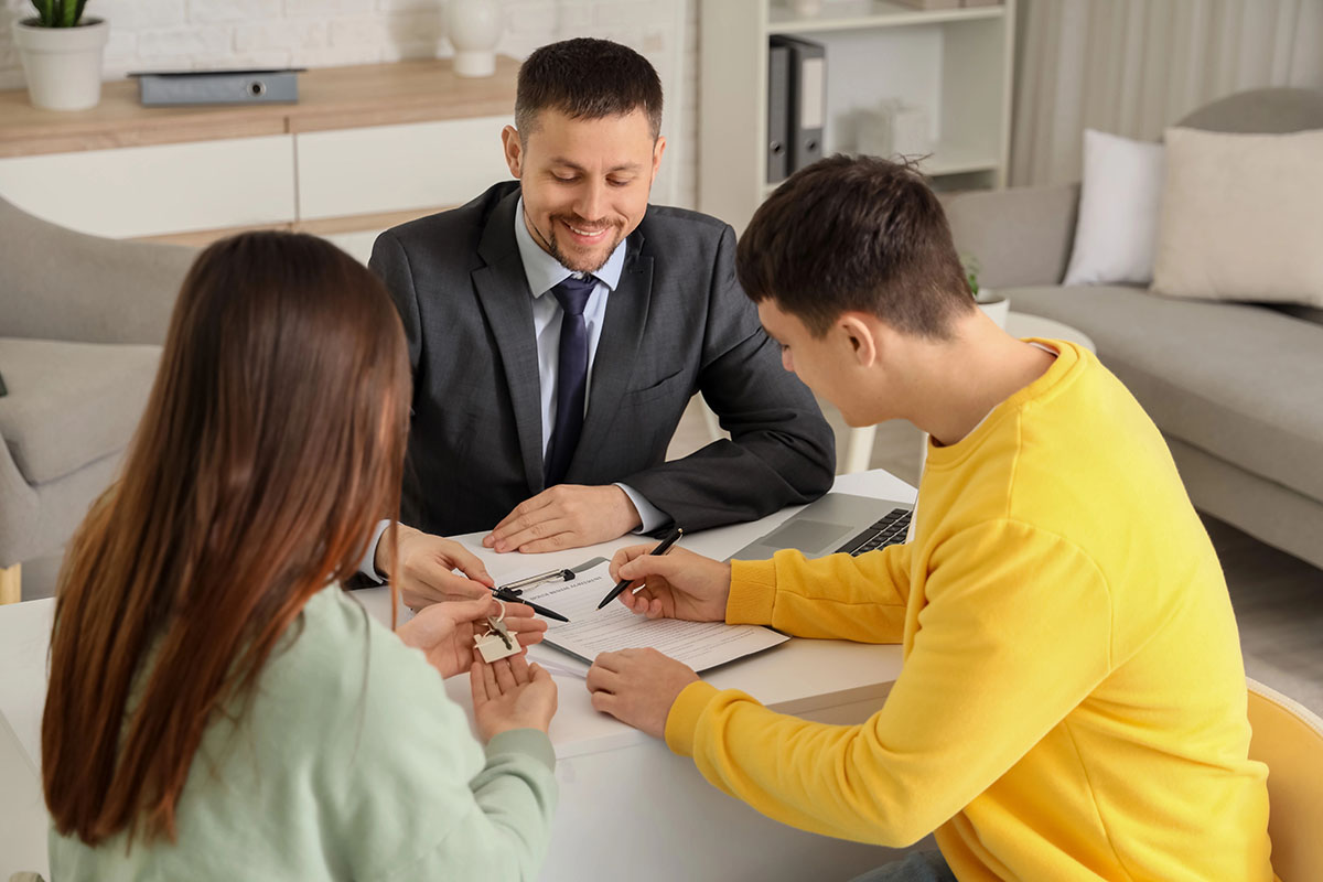 A professional man in a suit sits at a desk, smiling as he hands keys to a woman with long hair, who is wearing a green sweater. A young man in a yellow sweatshirt is looking at a document while holding a pen. A laptop is placed on the desk, and they appear to be discussing a transaction.