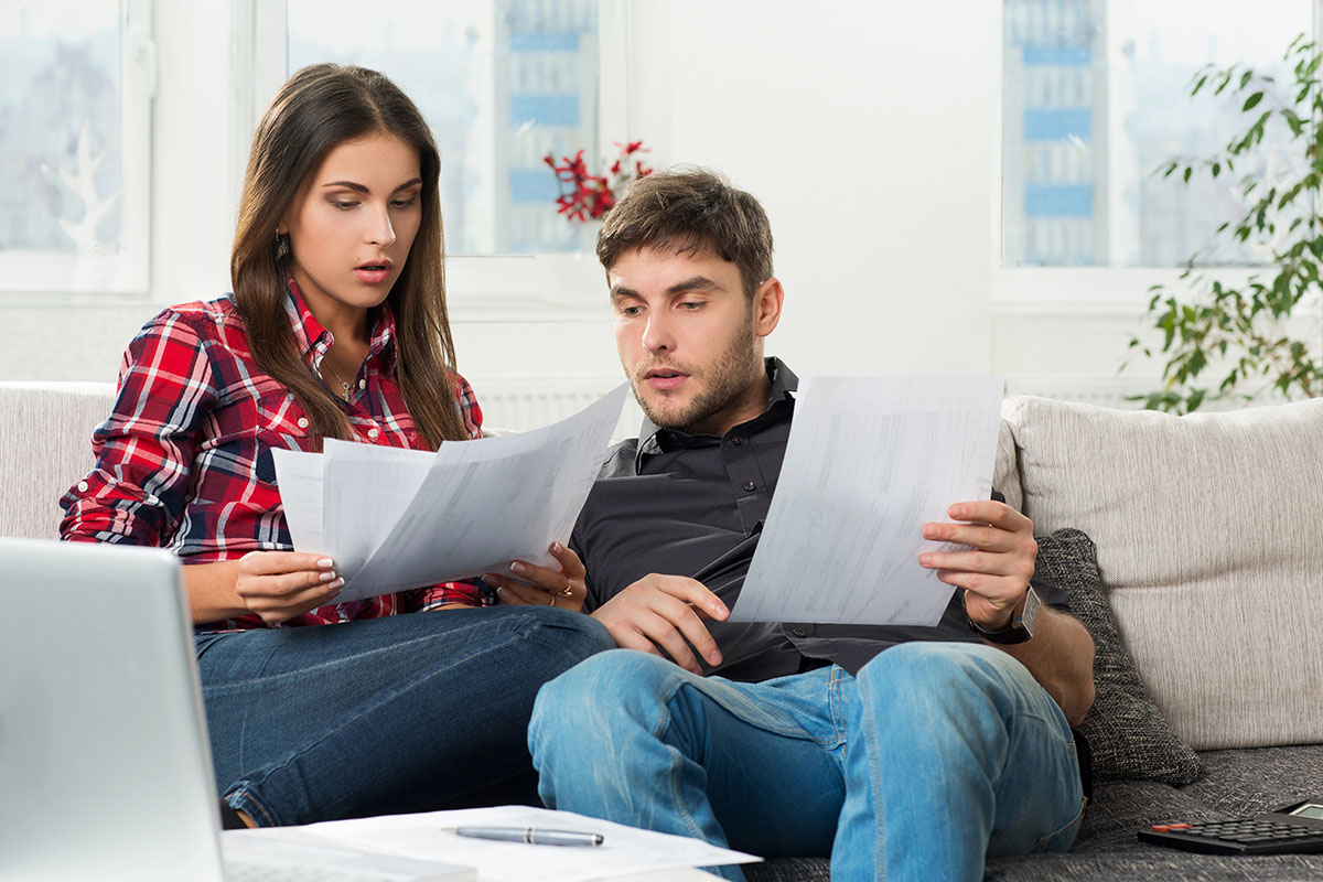 A man and a woman sit closely on a couch, looking intently at several sheets of paper in their hands, with a laptop and a pen on a table in front of them. Natural light filters through a window in the background.