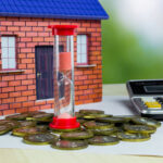 A decorative house model stands next to an hourglass filled with pink sand, surrounded by various coins and a calculator on a light-colored surface.