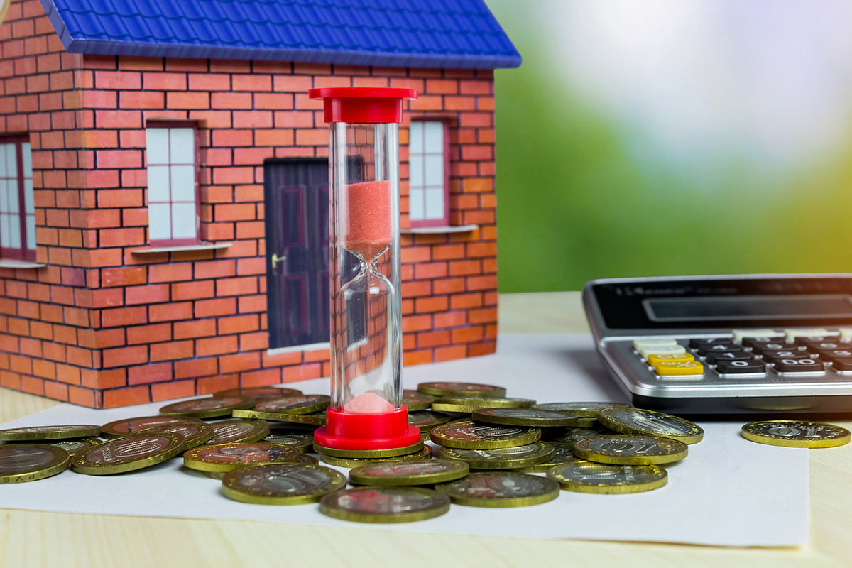 A decorative house model stands next to an hourglass filled with pink sand, surrounded by various coins and a calculator on a light-colored surface.