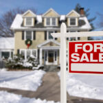 A "For Sale" sign stands in front of a house blanketed in snow, with a wreath on the door and a clear blue sky in the background.