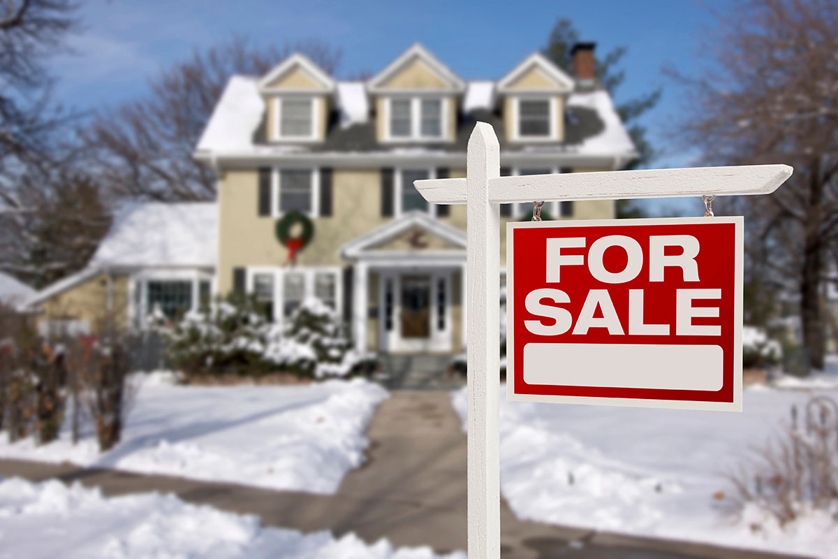 A "For Sale" sign stands in front of a house blanketed in snow, with a wreath on the door and a clear blue sky in the background.