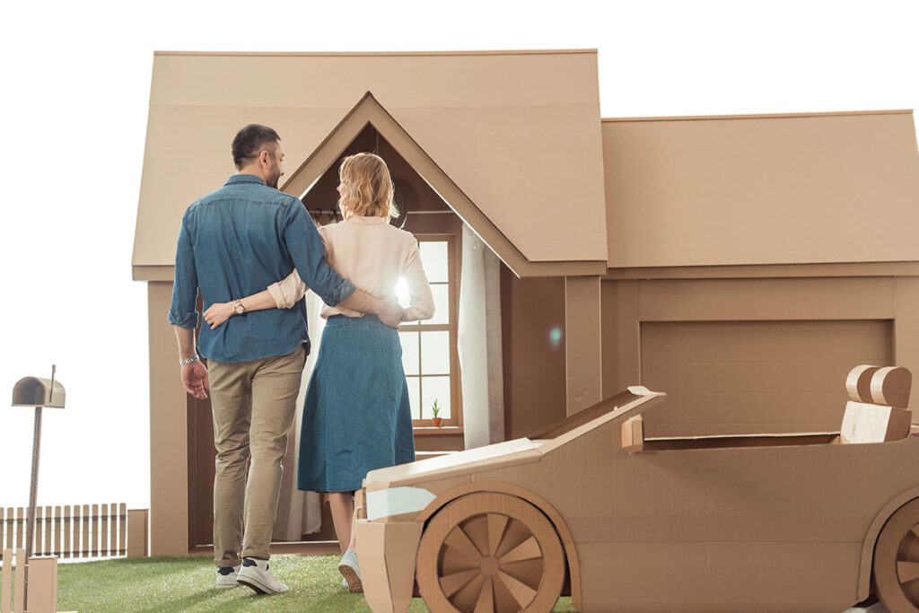 A couple stands together, viewing a large cardboard model of a house and a car, with the woman gently leaning against the man. They are positioned on a grassy surface.