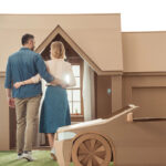 A couple stands together, viewing a large cardboard model of a house and a car, with the woman gently leaning against the man. They are positioned on a grassy surface.