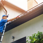 A person wearing a blue shirt and a cap is using a tool to clean the gutter while standing on a ladder beside a house.