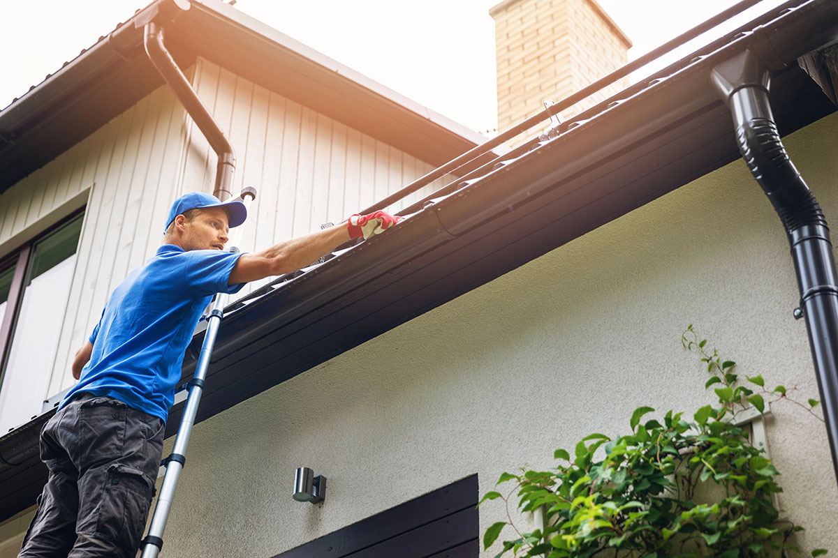 A person wearing a blue shirt and a cap is using a tool to clean the gutter while standing on a ladder beside a house.