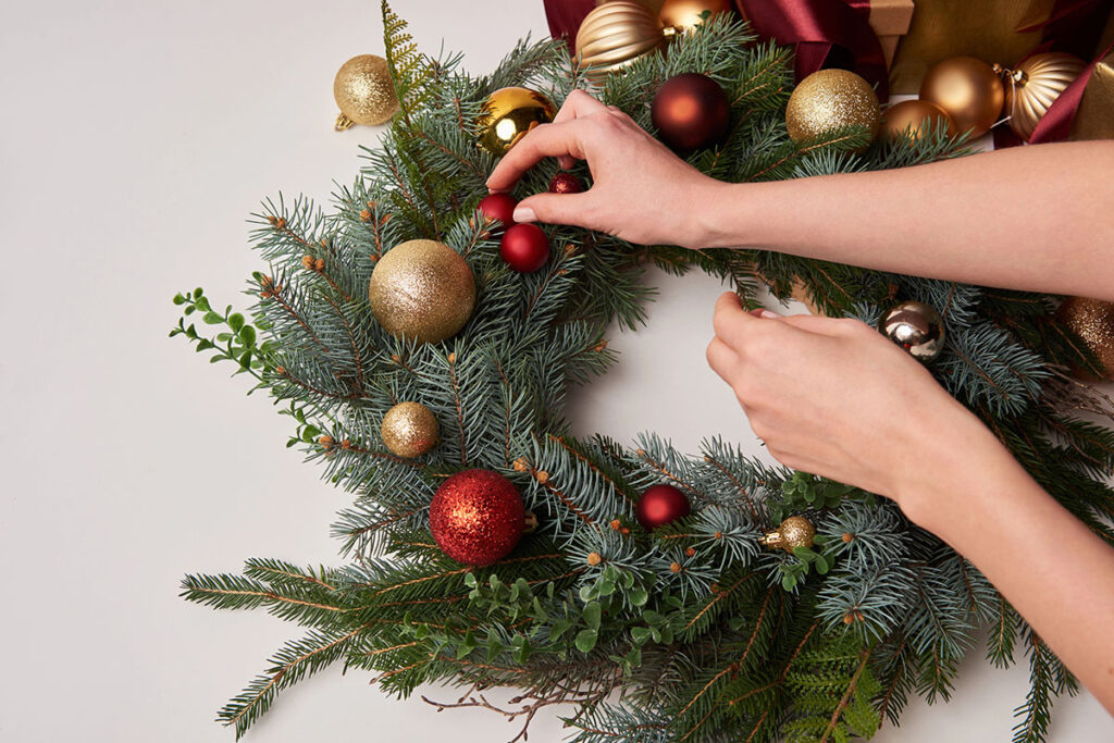 A person's hands adding red ornaments to a lush green Christmas wreath decorated with various colored balls, including gold and glittery red, against a neutral background.