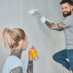 A man with tattoos is painting a wall with a roller while a woman with a ponytail and visible tattoos holds a glass of orange juice, watching him from the side.