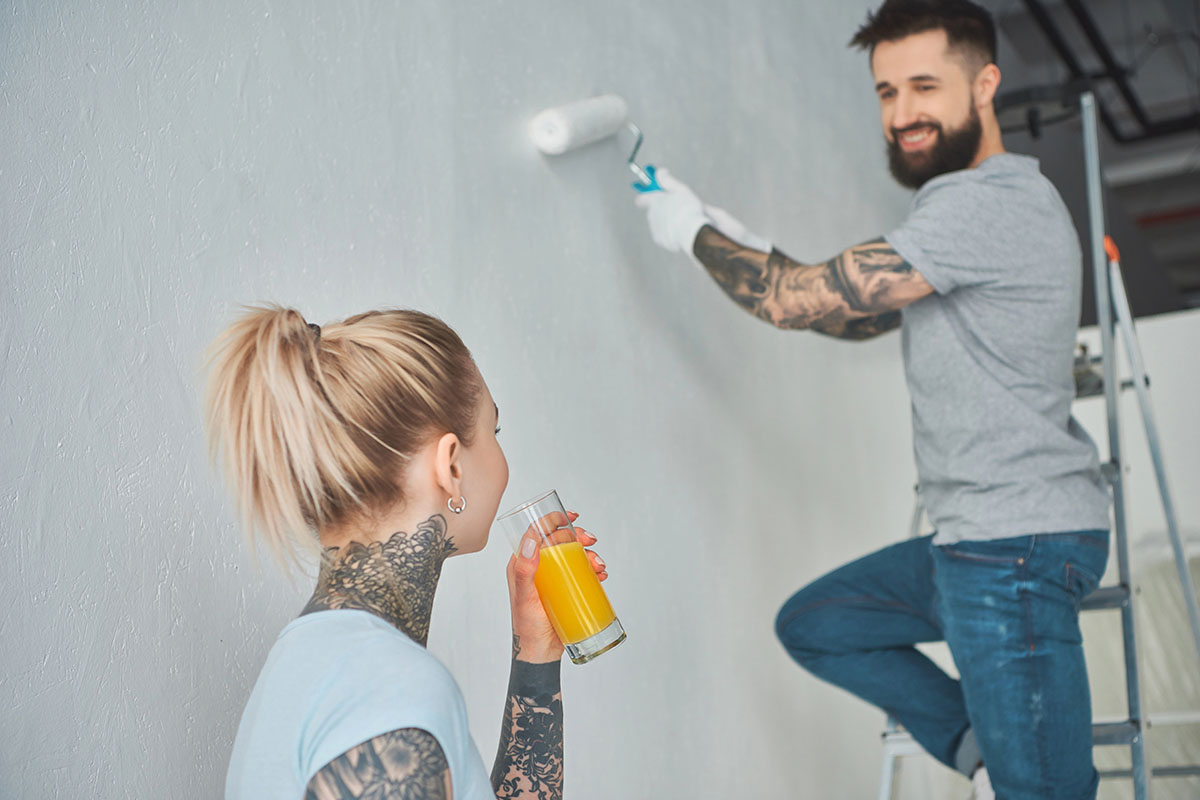 A man with tattoos is painting a wall with a roller while a woman with a ponytail and visible tattoos holds a glass of orange juice, watching him from the side.