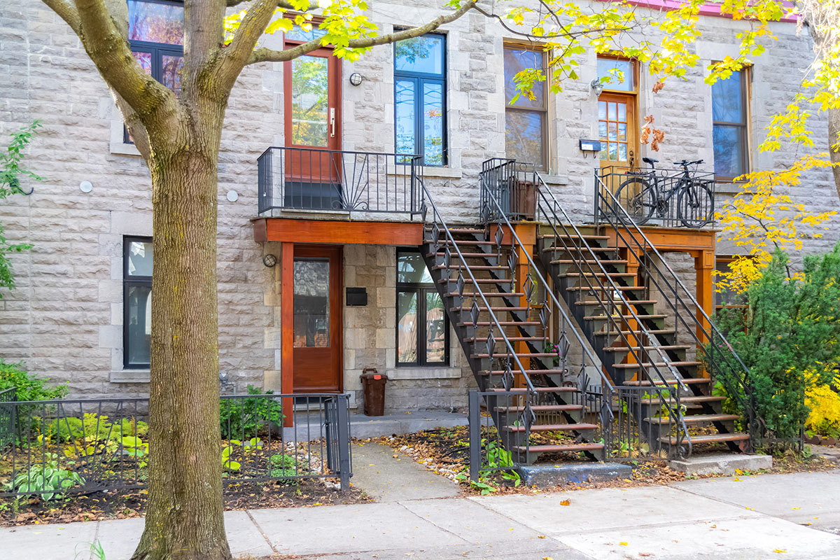 A stone building with two sets of outdoor stairs leading to wooden doors, surrounded by greenery and autumn foliage. One balcony features two bicycles.