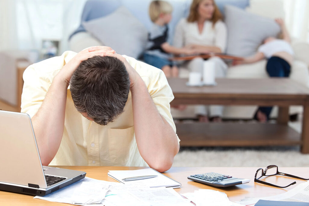 A man sits at a desk with his head in his hands, surrounded by papers and a laptop, showing signs of stress. In the background, two children and a woman are seated on a couch, engaged in conversation.