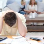 A man sits at a desk with his head in his hands, surrounded by papers and a laptop, showing signs of stress. In the background, two children and a woman are seated on a couch, engaged in conversation.