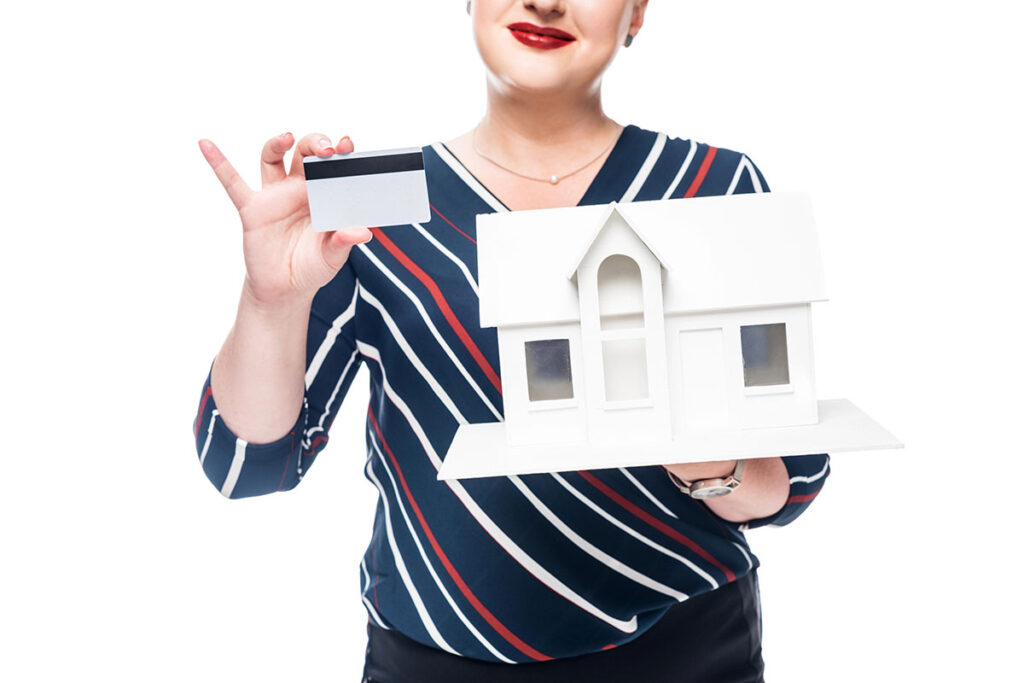 A smiling woman in a striped shirt holds a miniature white house in one hand and a credit card in the other, against a white background.