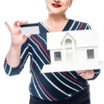 A smiling woman in a striped shirt holds a miniature white house in one hand and a credit card in the other, against a white background.