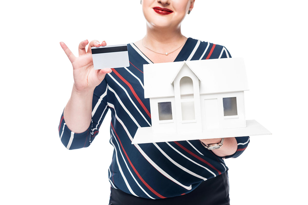 A smiling woman in a striped shirt holds a miniature white house in one hand and a credit card in the other, against a white background.