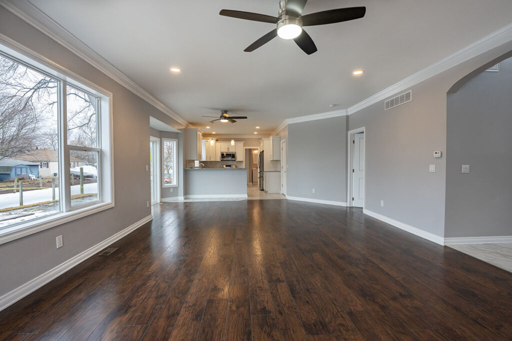 A spacious, modern living room featuring dark hardwood flooring, a ceiling fan, and large windows allowing natural light. The room leads to a contemporary kitchen with white cabinetry and stainless steel appliances, visible in the background.
