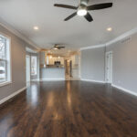 A spacious, modern living room featuring dark hardwood flooring, a ceiling fan, and large windows allowing natural light. The room leads to a contemporary kitchen with white cabinetry and stainless steel appliances, visible in the background.