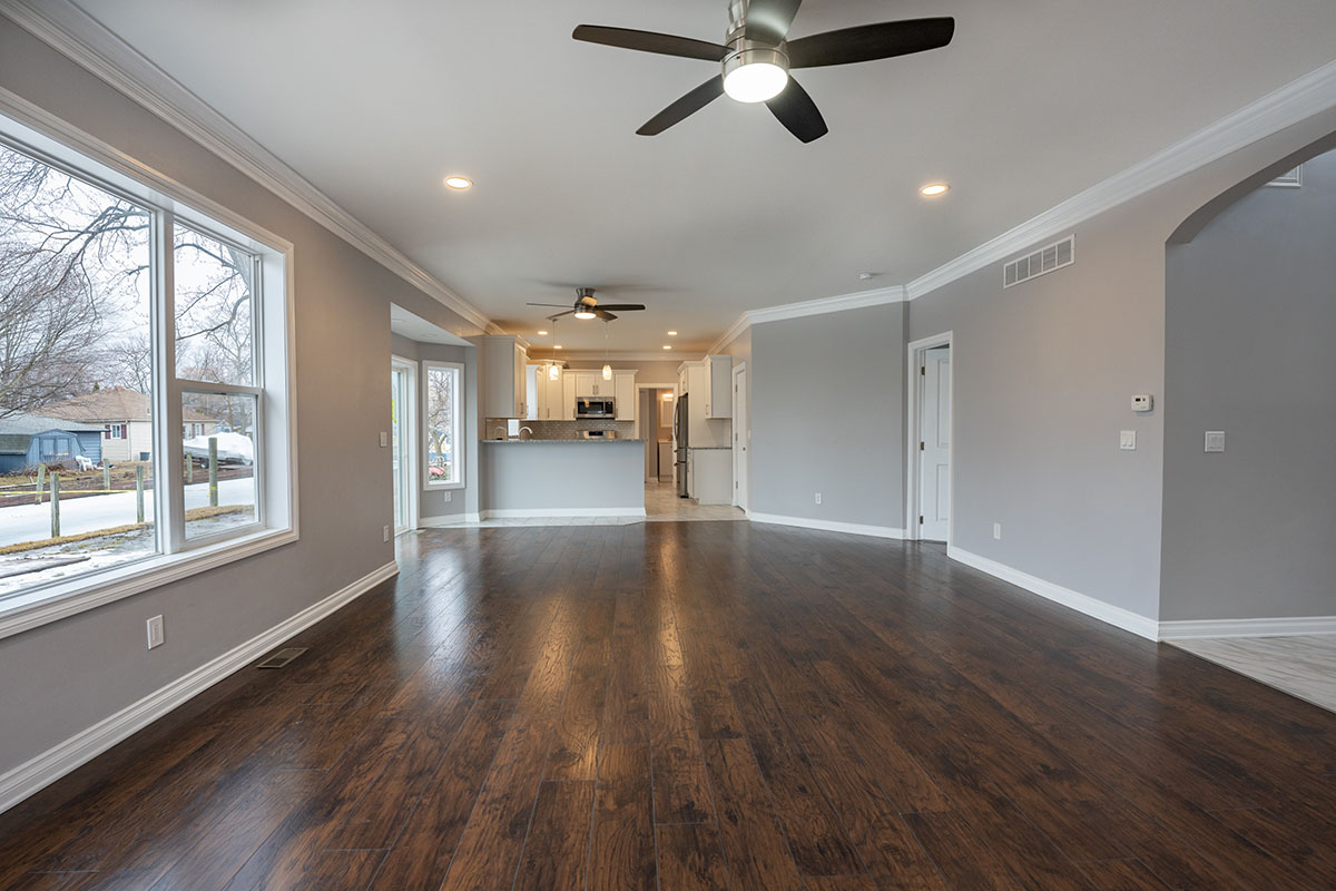 A spacious, modern living room featuring dark hardwood flooring, a ceiling fan, and large windows allowing natural light. The room leads to a contemporary kitchen with white cabinetry and stainless steel appliances, visible in the background.