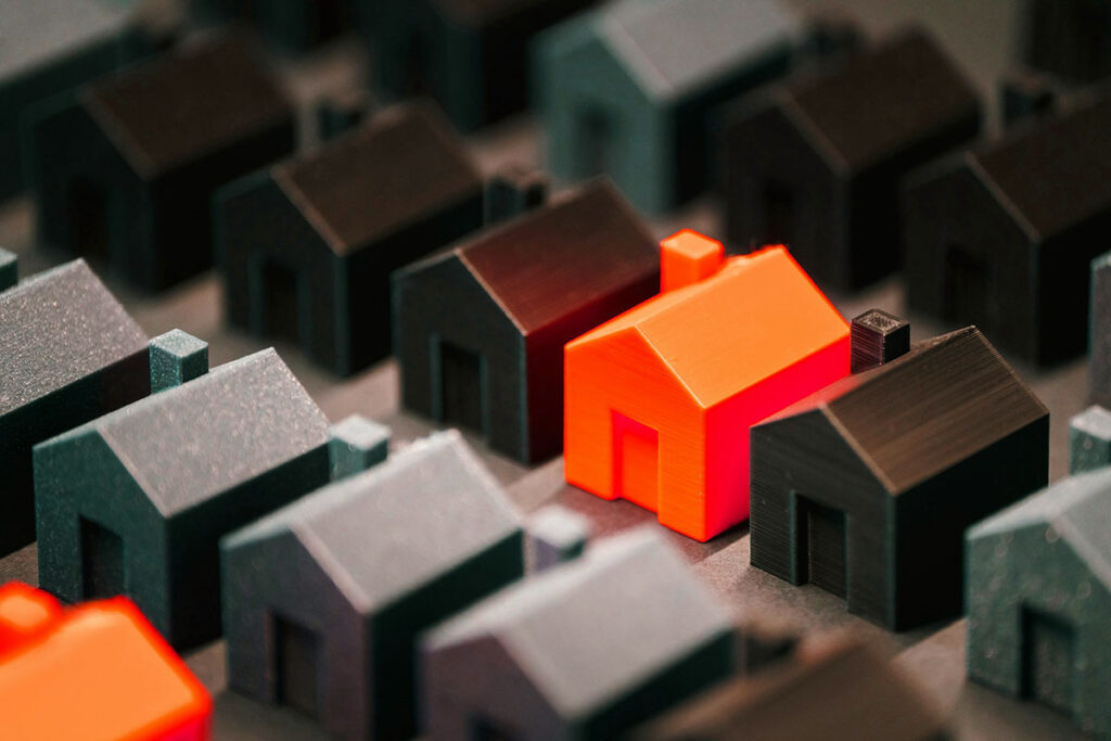 A bright orange house stands out among a cluster of dark-colored miniature houses, arranged on a surface.