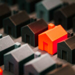 A bright orange house stands out among a cluster of dark-colored miniature houses, arranged on a surface.