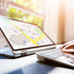 A close-up of a laptop displaying a map on the screen, with location markers visible, while a person's hands type on a keyboard. Bright sunlight filters through a window in the background.