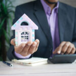 A business professional in a suit holds a colorful toy house in one hand while operating a calculator with the other, surrounded by a notepad and a pen on a wooden desk.